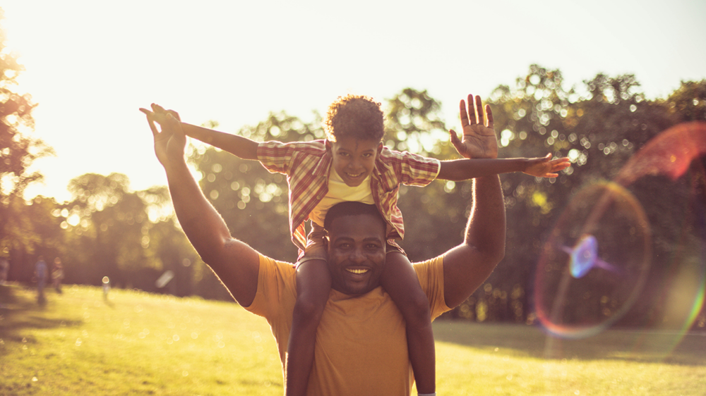 A man carries a child on his shoulders in a sunlit park. Both have their arms raised and are smiling.